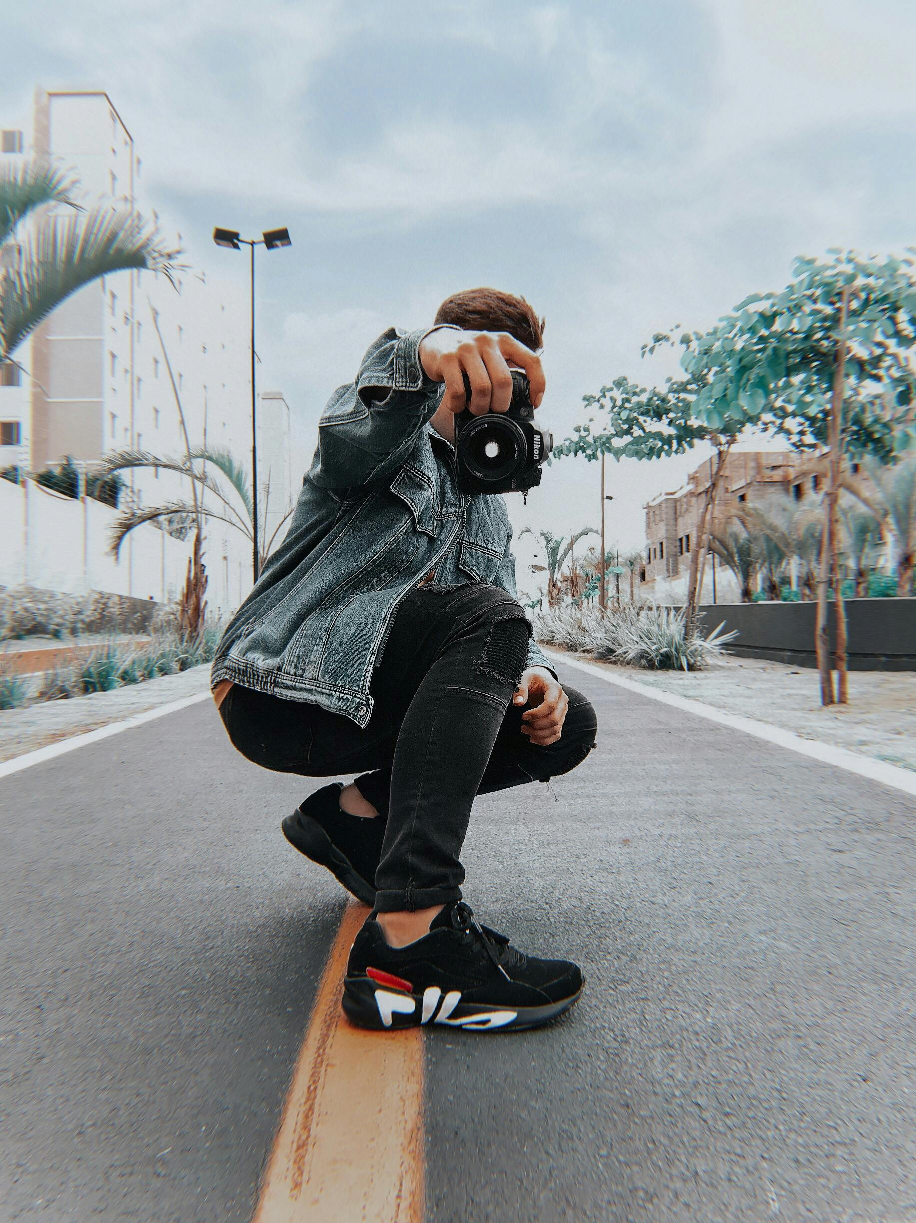 A male model stylishly poses with a camera on a city street in an urban setting.